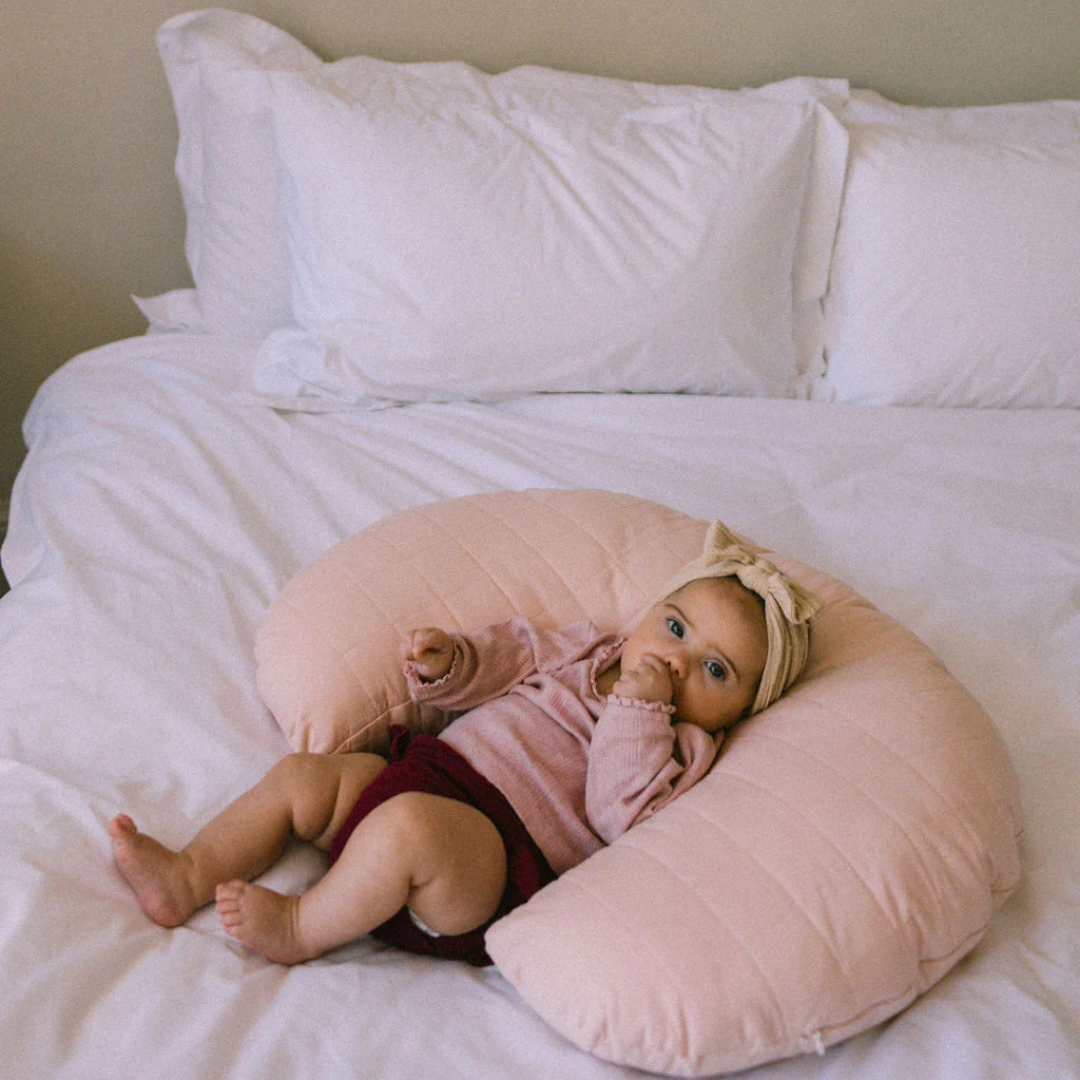Baby lying on a pink body pillow on a bed with white pillows and sheets.