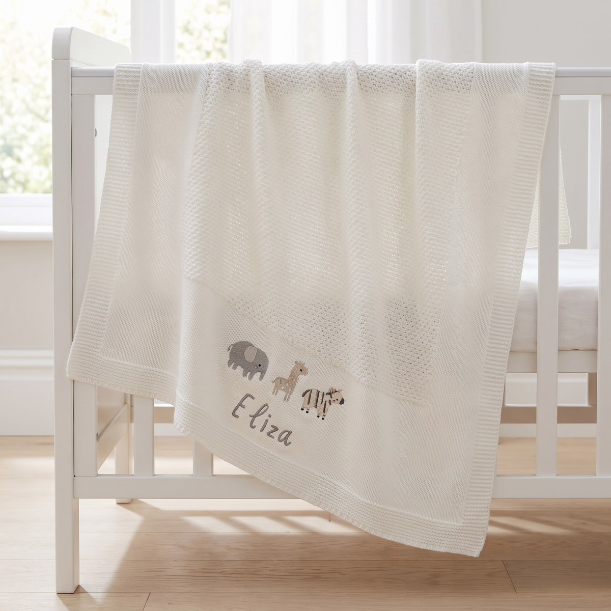 Personalised ivory knitted baby shawl blanket with embroidered name folded in a nursery